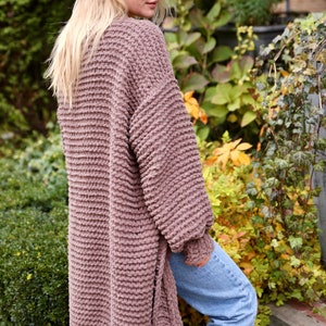 May include: A long, mauve knitted cardigan with a textured stitch pattern. The cardigan is open in the front and has long sleeves. The model is wearing the cardigan with blue jeans.