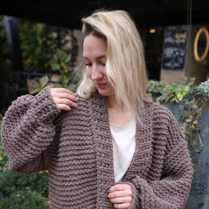 May include: A long, brown, chunky knit cardigan with a V-neck and long sleeves. The cardigan is made of a soft, fuzzy material and has a relaxed fit.