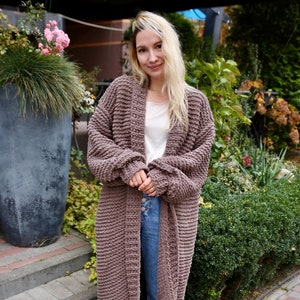May include: A long, brown knitted cardigan with a relaxed fit and a deep V-neck. The cardigan is worn over a white t-shirt and blue jeans.