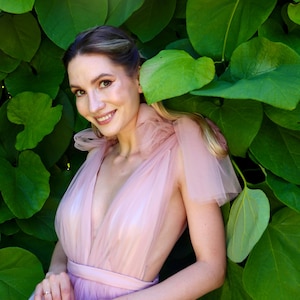 K&ouml;nnte beinhalten: Eine Person tr&auml;gt ein hellrosa T&uuml;llkleid mit tiefem Ausschnitt und taillierter Taille. Das Kleid hat einen mehrlagigen Rock und transparente &Auml;rmel. Sie l&auml;chelt vor einem Hintergrund aus gro&szlig;en gr&uuml;nen Bl&auml;ttern.