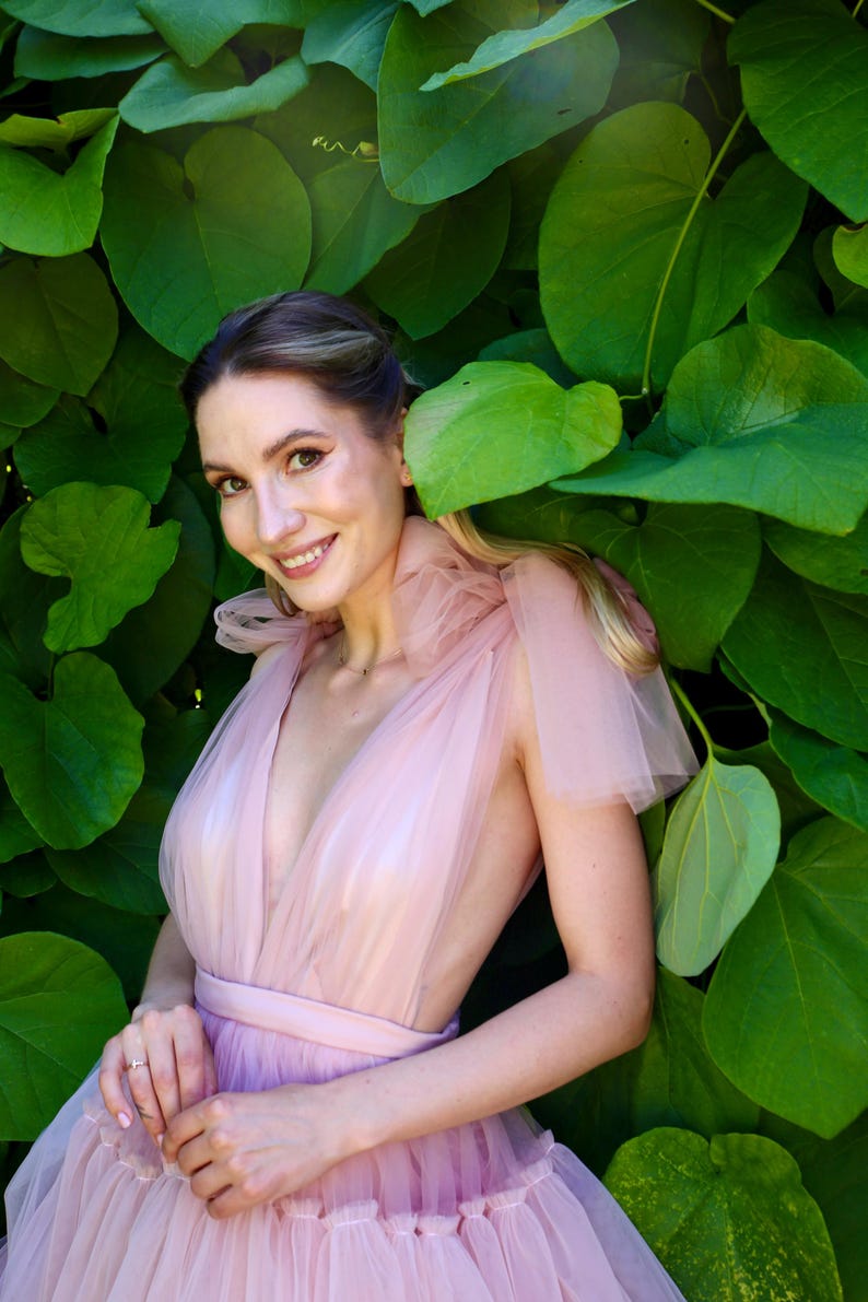 K&ouml;nnte beinhalten: Eine Person tr&auml;gt ein hellrosa T&uuml;llkleid mit tiefem Ausschnitt und taillierter Taille. Das Kleid hat einen mehrlagigen Rock und transparente &Auml;rmel. Sie l&auml;chelt vor einem Hintergrund aus gro&szlig;en gr&uuml;nen Bl&auml;ttern.