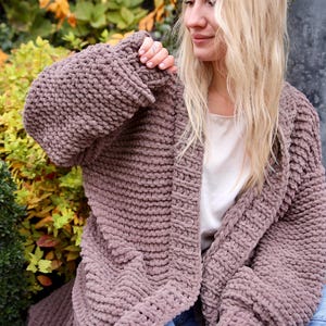 May include: A long, chunky knit cardigan in a mauve color. The cardigan is open in the front and has a relaxed fit. The model is wearing the cardigan over a white tank top and blue jeans.
