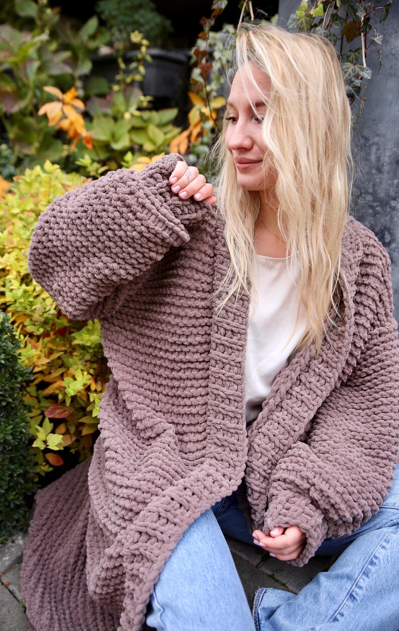 May include: A long, chunky knit cardigan in a mauve color. The cardigan is open in the front and has a relaxed fit. The model is wearing the cardigan over a white tank top and blue jeans.