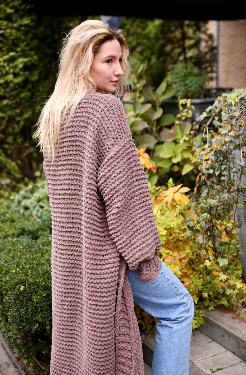 May include: A long, mauve knitted cardigan with a textured stitch pattern. The cardigan is open in the front and has long sleeves. The model is wearing the cardigan with blue jeans.