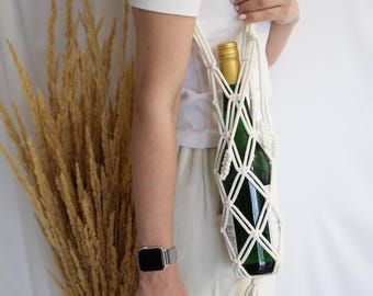 Puede incluir: Un porta botellas de macramé blanco con una borla que cuelga de la parte inferior. Una botella de vino está dentro del porta botellas.