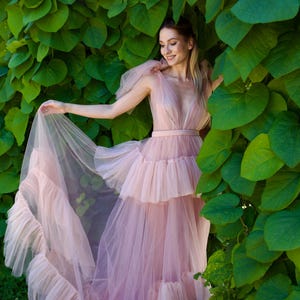K&ouml;nnte beinhalten: Ein hellrosa T&uuml;llkleid mit tiefem Ausschnitt und gestuftem Rock. Das Kleid hat R&uuml;schendetails und eine flie&szlig;ende Silhouette. Das Model steht vor einem Hintergrund aus &uuml;ppigen gr&uuml;nen Bl&auml;ttern, was eine weiche und romantische &Auml;sthetik erzeugt.