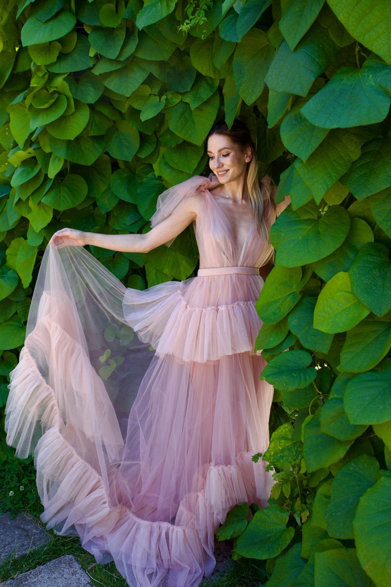 K&ouml;nnte beinhalten: Ein hellrosa T&uuml;llkleid mit tiefem Ausschnitt und gestuftem Rock. Das Kleid hat R&uuml;schendetails und eine flie&szlig;ende Silhouette. Das Model steht vor einem Hintergrund aus &uuml;ppigen gr&uuml;nen Bl&auml;ttern, was eine weiche und romantische &Auml;sthetik erzeugt.