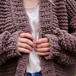 May include: A mauve knitted cardigan with a chunky knit texture. The cardigan is open in the front and has long sleeves.