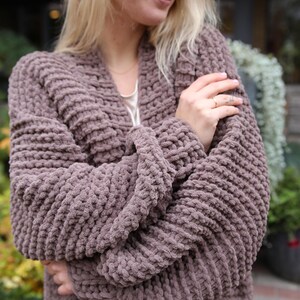 May include: A mauve knitted cardigan with a long, open front and a relaxed fit. The cardigan is made of a soft, chunky yarn and has a textured, waffle-like stitch.