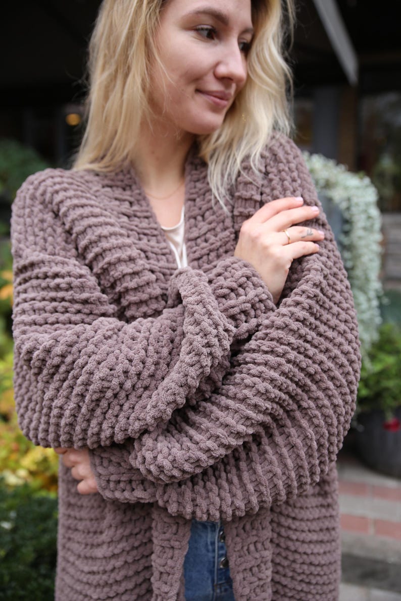 May include: A mauve knitted cardigan with a long, open front and a relaxed fit. The cardigan is made of a soft, chunky yarn and has a textured, waffle-like stitch.