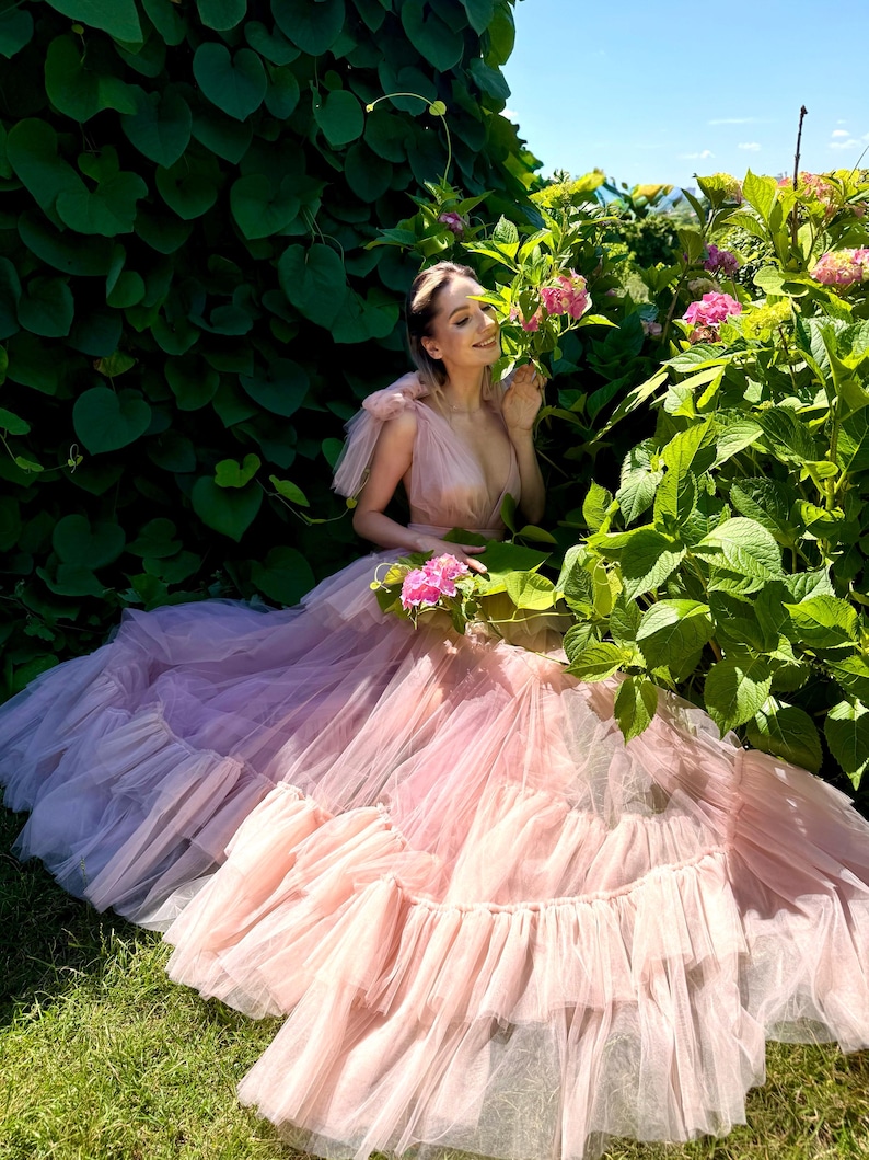 K&ouml;nnte beinhalten: Ein hellrosa T&uuml;llkleid mit einem mehrlagigen Rock und R&uuml;schensaum. Das Kleid hat einen tiefen Ausschnitt und kurze, ger&uuml;schte &Auml;rmel. Das Kleid wird im Freien getragen, umgeben von Gr&uuml;n und rosa Blumen.