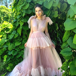 K&ouml;nnte beinhalten: Ein hellrosa T&uuml;llkleid mit tiefem Ausschnitt und mehrlagigem Rock. Das Kleid hat ein eng anliegendes Oberteil und einen flie&szlig;enden, gestuften Rock. Das Kleid wird im Freien getragen, umgeben von &uuml;ppigem gr&uuml;nem Laub. Das Kleid hat ein zartes, &auml;therisches Aussehen.