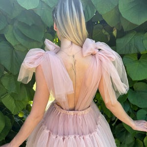 K&ouml;nnte beinhalten: Hellrosa T&uuml;llkleid mit tiefem R&uuml;ckenausschnitt und geschichtetem Rock. Das Kleid hat gro&szlig;e Schleifen an den Schultern und eine taillierte Taille. Der Hintergrund ist gr&uuml;nes Laub.