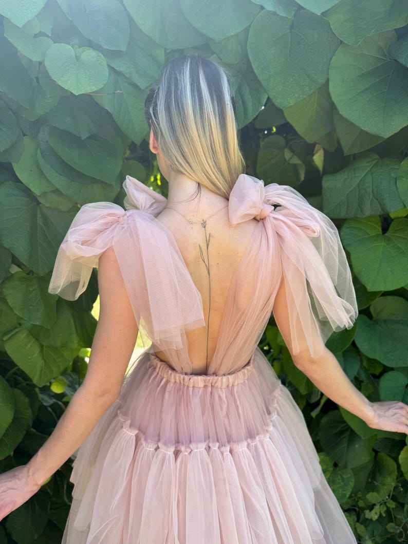 K&ouml;nnte beinhalten: Hellrosa T&uuml;llkleid mit tiefem R&uuml;ckenausschnitt und geschichtetem Rock. Das Kleid hat gro&szlig;e Schleifen an den Schultern und eine taillierte Taille. Der Hintergrund ist gr&uuml;nes Laub.