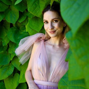 K&ouml;nnte beinhalten: Eine Frau in einem hellrosa T&uuml;llkleid mit tiefem Ausschnitt und passendem G&uuml;rtel. Das Kleid hat einen geschichteten Rock und transparente, ger&uuml;schte &Auml;rmel. Der Hintergrund ist mit &uuml;ppigen gr&uuml;nen Bl&auml;ttern gef&uuml;llt.