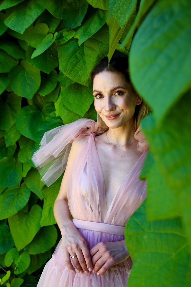 K&ouml;nnte beinhalten: Eine Frau in einem hellrosa T&uuml;llkleid mit tiefem Ausschnitt und passendem G&uuml;rtel. Das Kleid hat einen geschichteten Rock und transparente, ger&uuml;schte &Auml;rmel. Der Hintergrund ist mit &uuml;ppigen gr&uuml;nen Bl&auml;ttern gef&uuml;llt.