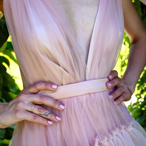 K&ouml;nnte beinhalten: Hellrosa T&uuml;llkleid mit tiefem V-Ausschnitt und taillierter Taille. Das Kleid hat einen mehrlagigen Rock mit R&uuml;schensaum. Das Model tr&auml;gt eine goldene Halskette und einen Ring. Der Hintergrund ist ein verschwommenes Gr&uuml;n.