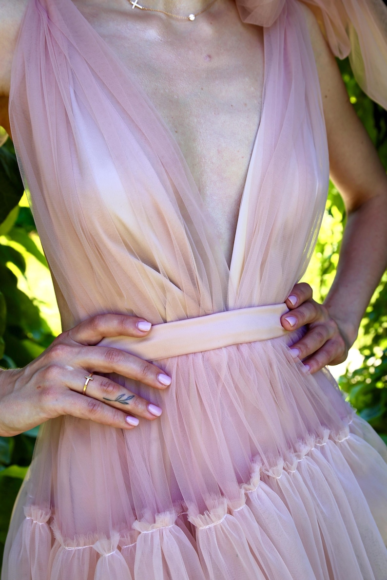 K&ouml;nnte beinhalten: Hellrosa T&uuml;llkleid mit tiefem V-Ausschnitt und taillierter Taille. Das Kleid hat einen mehrlagigen Rock mit R&uuml;schensaum. Das Model tr&auml;gt eine goldene Halskette und einen Ring. Der Hintergrund ist ein verschwommenes Gr&uuml;n.