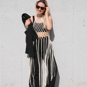 以下が含まれることがあります： 白いフリンジの付いた白いクロシェトップと黒いパンツを着用した女性。彼女はサングラスをかけており、黒いブレザーを持っています。