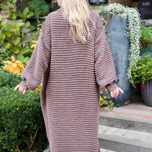 May include: A long, brown, knitted cardigan with a loose fit and a ribbed texture. The cardigan is shown from the back and is worn by a person with long blonde hair.