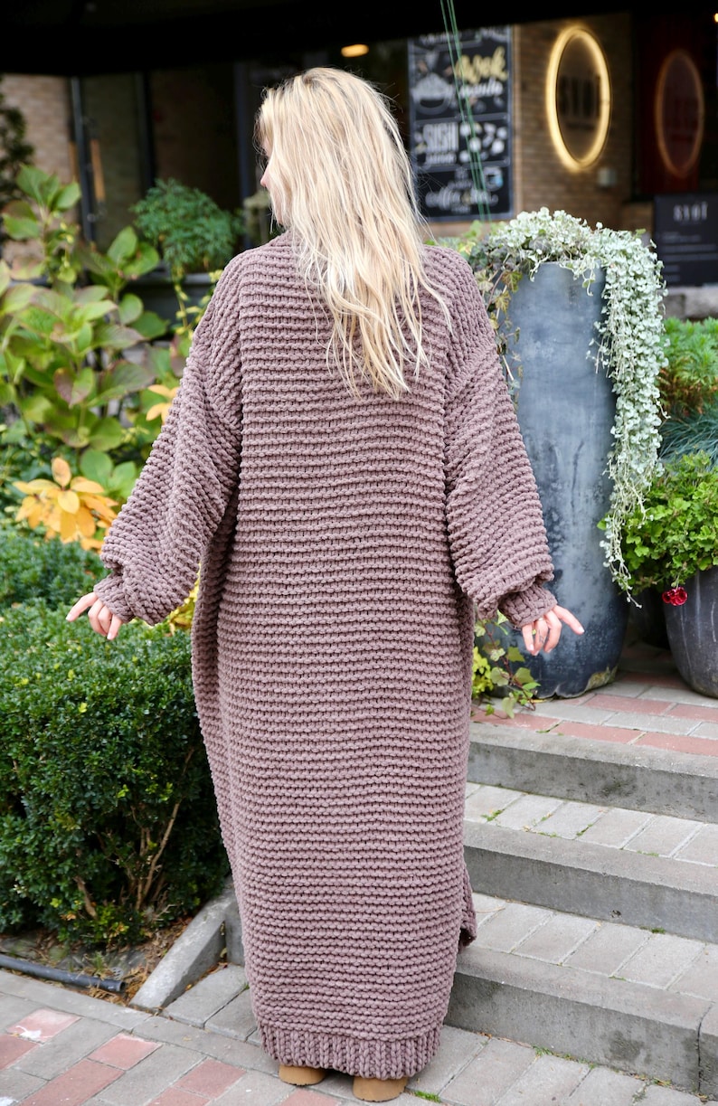 May include: A long, brown, knitted cardigan with a loose fit and a ribbed texture. The cardigan is shown from the back and is worn by a person with long blonde hair.