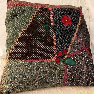 May include: A patchwork pillow with a variety of patterned fabrics in shades of green, brown, and black. The pillow features embroidered details, including a red flower, green leaves, and a stitched bow.