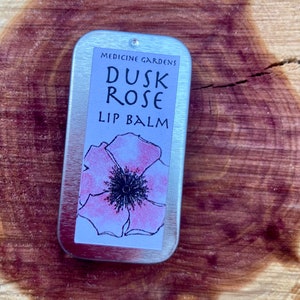 Puede incluir: Una lata de plata con una etiqueta blanca que dice "Medicine Gardens Dusk Rose Lip Balm". La etiqueta presenta una flor rosa con un centro negro.