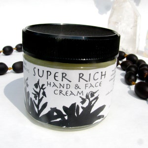 May include: A glass jar with a black lid filled with a light green cream. The jar has a white label with black text that reads "Super Rich Hand & Face Cream".