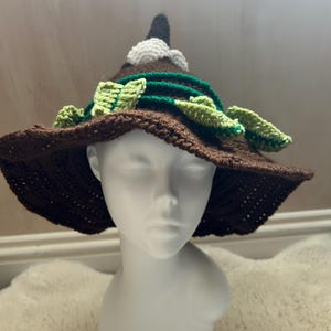 May include: A brown crochet hat with a wide brim, adorned with green leaf accents and a white flower. The hat has a pointed crown and is displayed on a white mannequin head. The hat's design is reminiscent of a witch's hat.