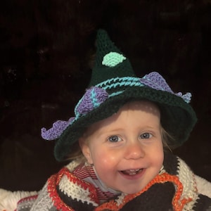 May include: A dark green crocheted pointed hat featuring a light blue accent band and purple decorative elements. The hat is paired with a multi-colored crocheted wrap in shades of white, gray, black, and orange with metallic gold threading.