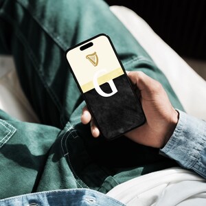 May include: A black smartphone with a white and gold case. The case has a harp logo and the letter "G" on it.