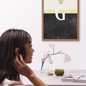 May include: A framed print with a black and white abstract design. The design features a gold harp symbol and the letter "G" in white. The print is hanging on a white wall.