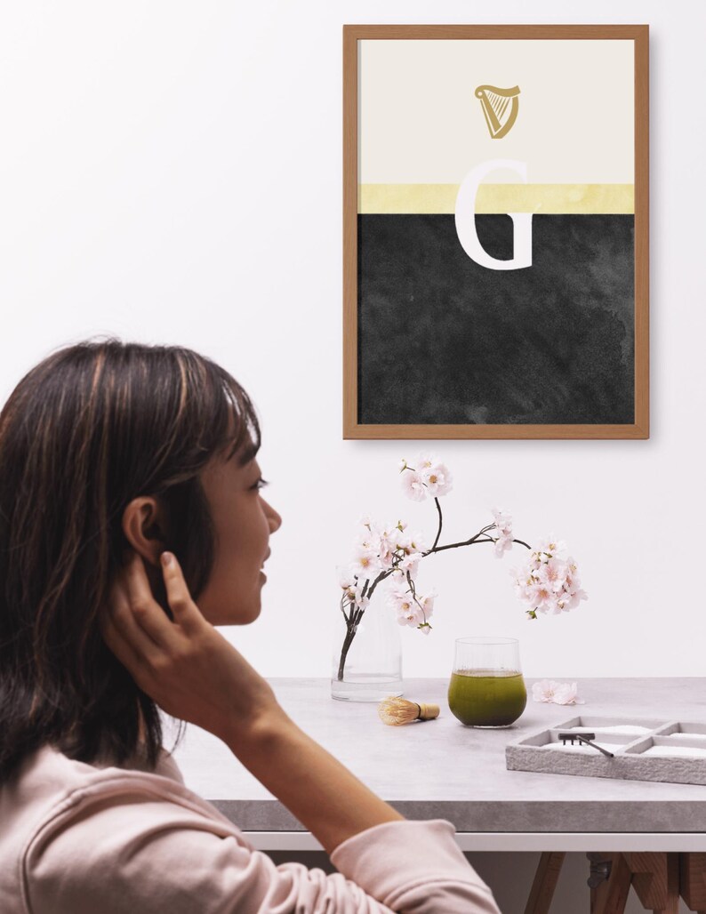 May include: A framed print with a black and white abstract design. The design features a gold harp symbol and the letter "G" in white. The print is hanging on a white wall.
