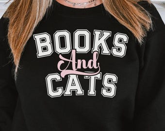 May include: Black sweatshirt with the text "BOOKS And CATS" in white block letters with a pink "And". The sweatshirt is a crew neck style and is being worn by a person. The background is a kitchen.