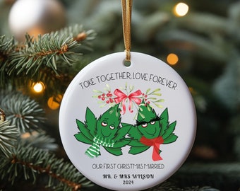 Może przedstawiać: Okrągła, biała ozdoba choinkowa ze złotym sznurkiem. Ozdoba przedstawia dwa kreskówkowe liście konopi z szalikami i napis "Toke Together, Love Forever". Poniżej napis "Our First Christmas Married" i "Mr. & Mrs. Wilson 2024."