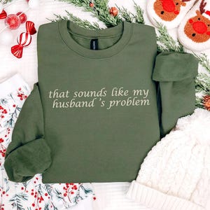 Może przedstawiać: Oliwkowa bluza z napisem "that sounds like my husband's problem" w białym skrypcie. Obraz zawiera świąteczne dekoracje, takie jak cukierki, zieleń i biała dzianinowa czapka.