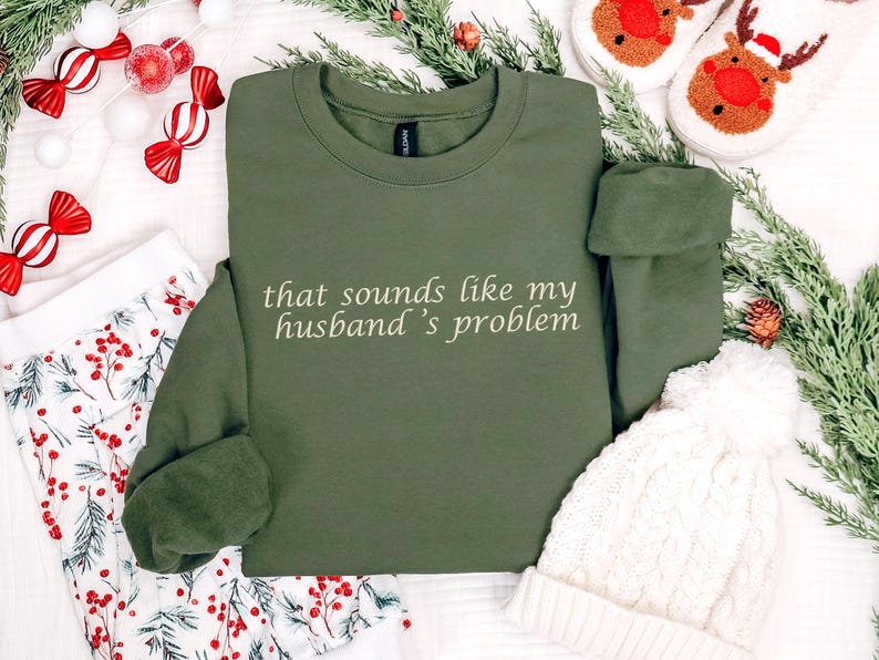 Może przedstawiać: Oliwkowa bluza z napisem "that sounds like my husband's problem" w białym skrypcie. Obraz zawiera świąteczne dekoracje, takie jak cukierki, zieleń i biała dzianinowa czapka.