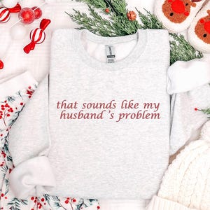 Może przedstawiać: Jasnoszara bluza z napisem "that sounds like my husband's problem" w bordowych literach. Obraz zawiera świąteczną piżamę, białą czapkę i kapcie z reniferami.