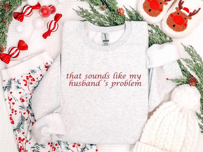 Może przedstawiać: Jasnoszara bluza z napisem "that sounds like my husband's problem" w bordowych literach. Obraz zawiera świąteczną piżamę, białą czapkę i kapcie z reniferami.