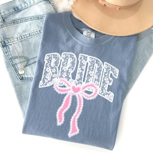 May include: A blue t-shirt with the word "BRIDE" in white floral lace lettering and a pink bow graphic. The shirt is paired with light wash jeans and a tan hat.