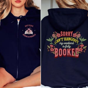 以下が含まれることがあります： ネイビーブルーのジップアップパーカー。フロントには花が描かれた開いた本と「QUIET PLEASE READING IN PROGRESS」の文字がプリントされています。バックには花柄のデザインと「SORRY CAN'T HANGOUT my weekend is fully BOOKED」の文字が施されています。
