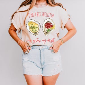 May include: A light beige t-shirt with a graphic of two oysters. The text "I'm a bit shellfish, my oysters, my delight" is printed on the shirt in red.