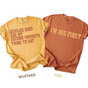 May include: Two short-sleeve crew neck t-shirts. The mustard yellow shirt features brown text reading "DEVILED EGGS ARE MY SECOND FAVORITE THING TO EAT". The yam orange shirt features brown text reading "I'M HIS FIRST". Both shirts have a casual fit.