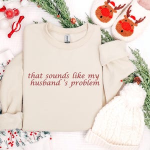 Może przedstawiać: Kremowa bluza z napisem "that sounds like my husband's problem" w bordowym skrypcie. Obraz zawiera kapcie z reniferami, białą dzianinową czapkę i świąteczne dekoracje.