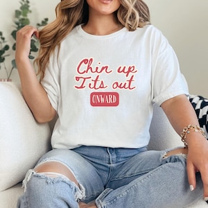 May include: White t-shirt with the text "Chin up Its out" in red lettering and a red rectangle with the word "ONWARD" inside. The person is wearing blue jeans.