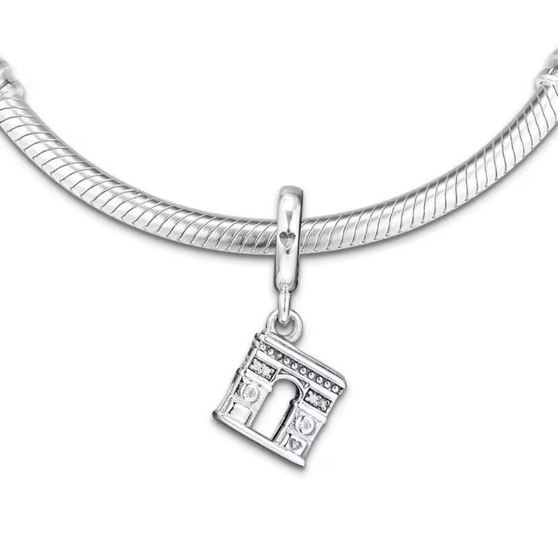 May include: Silver charm in the shape of the Arc de Triomphe, a famous landmark in Paris, France. The charm has a detailed design and is perfect for adding a touch of Parisian style to a bracelet or necklace.