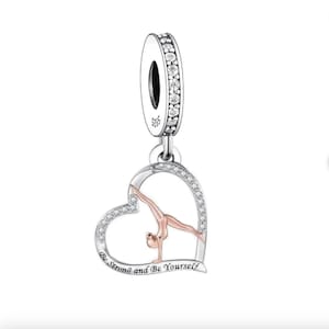 Könnte beinhalten: Ein silberner Charm-Anhänger mit einem herzförmigen Anhänger, der eine Turnerin im Handstand zeigt. Das Herz ist mit klaren Kristallen umrandet und trägt die Aufschrift "Be Strong and Be Yourself". Der Charm hat eine silberne Schlaufe mit klaren Kristallen.