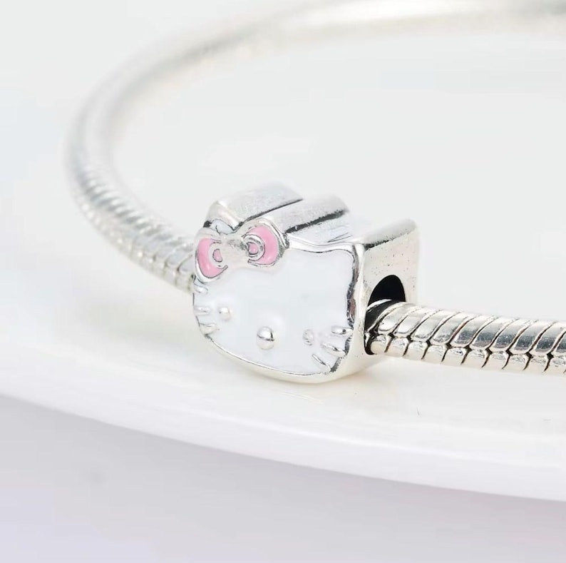 May include: A silver charm bracelet with a white Hello Kitty charm with pink accents. The charm has a bow and a face with black eyes and whiskers.