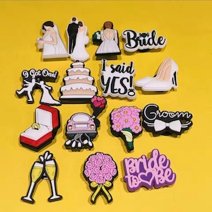 Op de afbeelding: Een verzameling bruiloft-thema bedels en versieringen. Inclusief bruid- en bruidegomfiguren, een bruidstaart, een ring in een doos, een auto, boeketten, champagneglazen en tekst met "Bride", "Groom", "I said YES!", en "Bride to be".