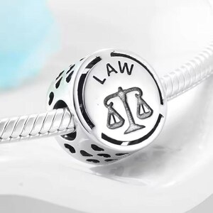 Könnte beinhalten: Silberner Anhänger mit rundem Design, das das Wort "LAW" und eine Justiwaage zeigt. Der Anhänger hat ein detailliertes, durchbrochenes Design.